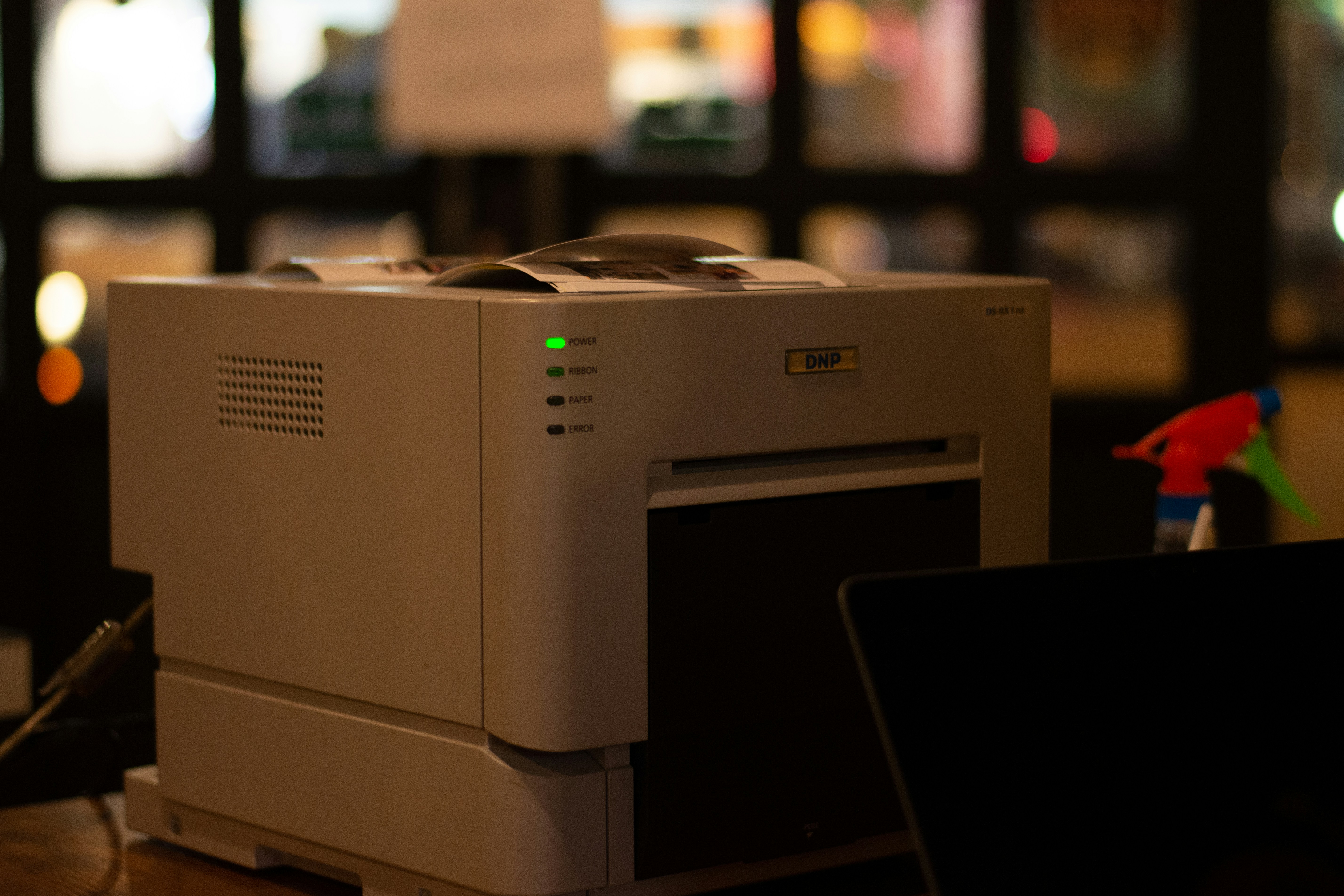 A DNP photo printer on a desk, power and ribbon lights glowing green in a dark room